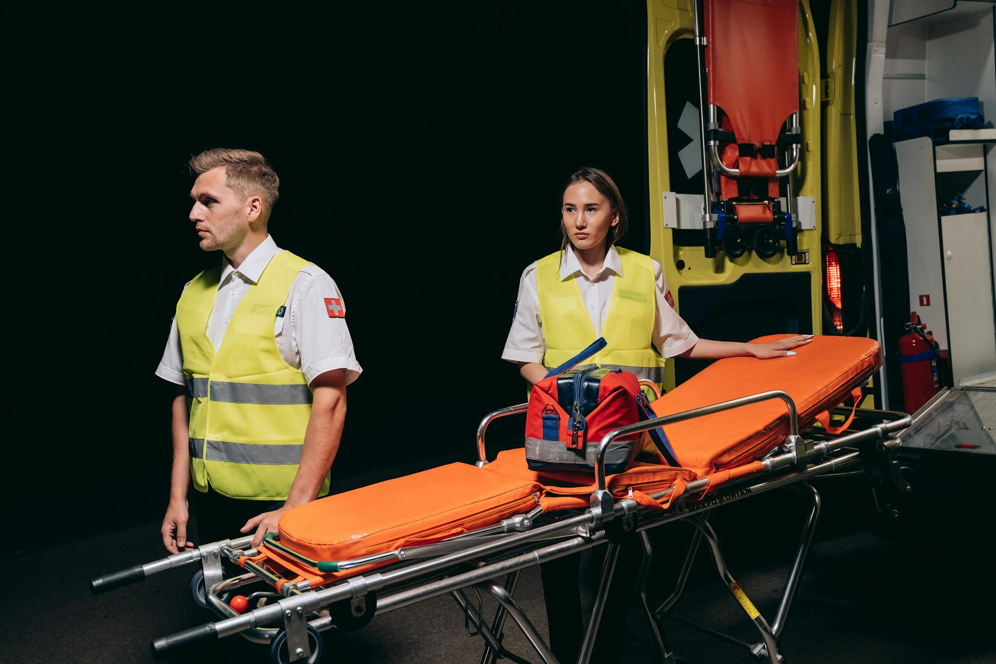 Two paramedics preparing medical stretcher by ambulance at night.